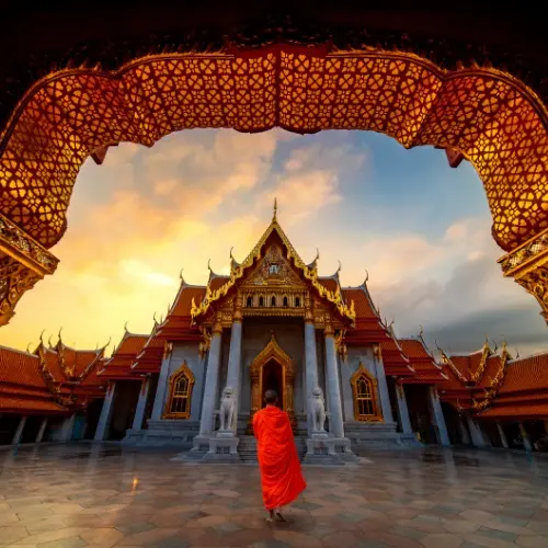 Buddhist monk in orange robes walking toward the Marble Temple (Wat Benchamabophit) in Bangkok at sunrise, framed by ornate golden archway